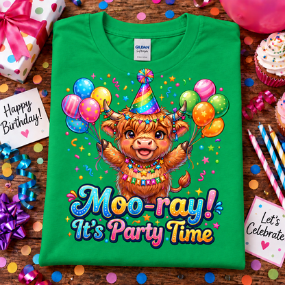 A irish green Gildan Comfort short-sleeve t-shirt laid flat on a wooden surface, decorated with a colorful cartoon Highland cow holding balloons and the text 'Moo-ray! It's Party Time.' Birthday-themed props like cupcakes, confetti, ribbons, and cards surround the shirt.