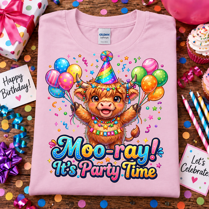 A folded light pink Gildan Comfort T-shirt is displayed on a wooden surface surrounded by birthday party props, including confetti, cupcakes, balloons, ribbons, and cards reading 'Happy Birthday!' and 'Let's Celebrate.' The shirt features a colorful cartoon Highland cow wearing a party hat and holding balloons, with the text 'Moo-ray! It's Party Time.