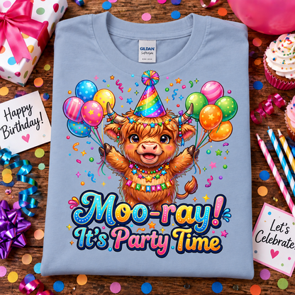 A light blue Gildan Comfort short-sleeve t-shirt laid flat on a wooden surface, decorated with a colorful cartoon Highland cow character holding balloons and the text 'Moo-ray! It's Party Time.' Surrounding the shirt are birthday party props including wrapped gifts, cupcakes, confetti, streamers, and small signs.