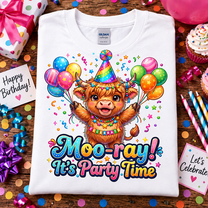 A folded white Gildan Comfort T-shirt is displayed on a wooden surface surrounded by birthday party props, including confetti, wrapped gifts, cupcakes, and party hats. The shirt features a colorful cartoon illustration of a Highland cow wearing a party hat and necklace, holding balloons, with the text 'Moo-ray! It's Party Time'.
