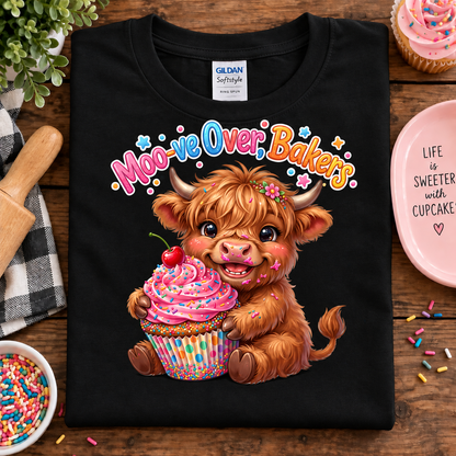 A black Gildan Softstyle t-shirt laid flat on a wooden table, featuring a colorful graphic of a fluffy brown highland cow holding a pink cupcake with a cherry. The text above reads 'Moo-ve Over Bakesh' in playful multicolored lettering.