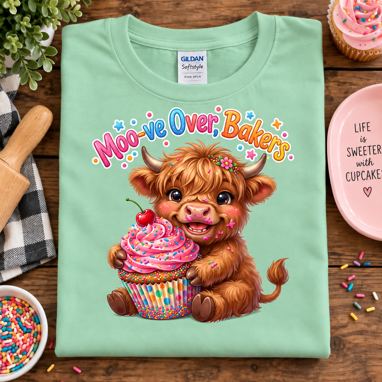 A mint green Gildan Sofstyle t-shirt laid flat on a wooden table, featuring a cute graphic of a fluffy brown highland cow holding a pink frosted cupcake with a cherry on top. The text 'Moo-ve Over, Bakers' is written above the cow in a playful multicolor font.