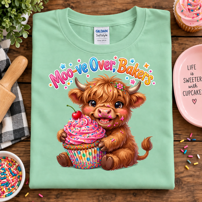A mint green Gildan Sofstyle t-shirt laid flat on a wooden table, featuring a cute graphic of a fluffy brown highland cow holding a pink frosted cupcake with a cherry on top. The text 'Moo-ve Over, Bakers' is written above the cow in a playful multicolor font.