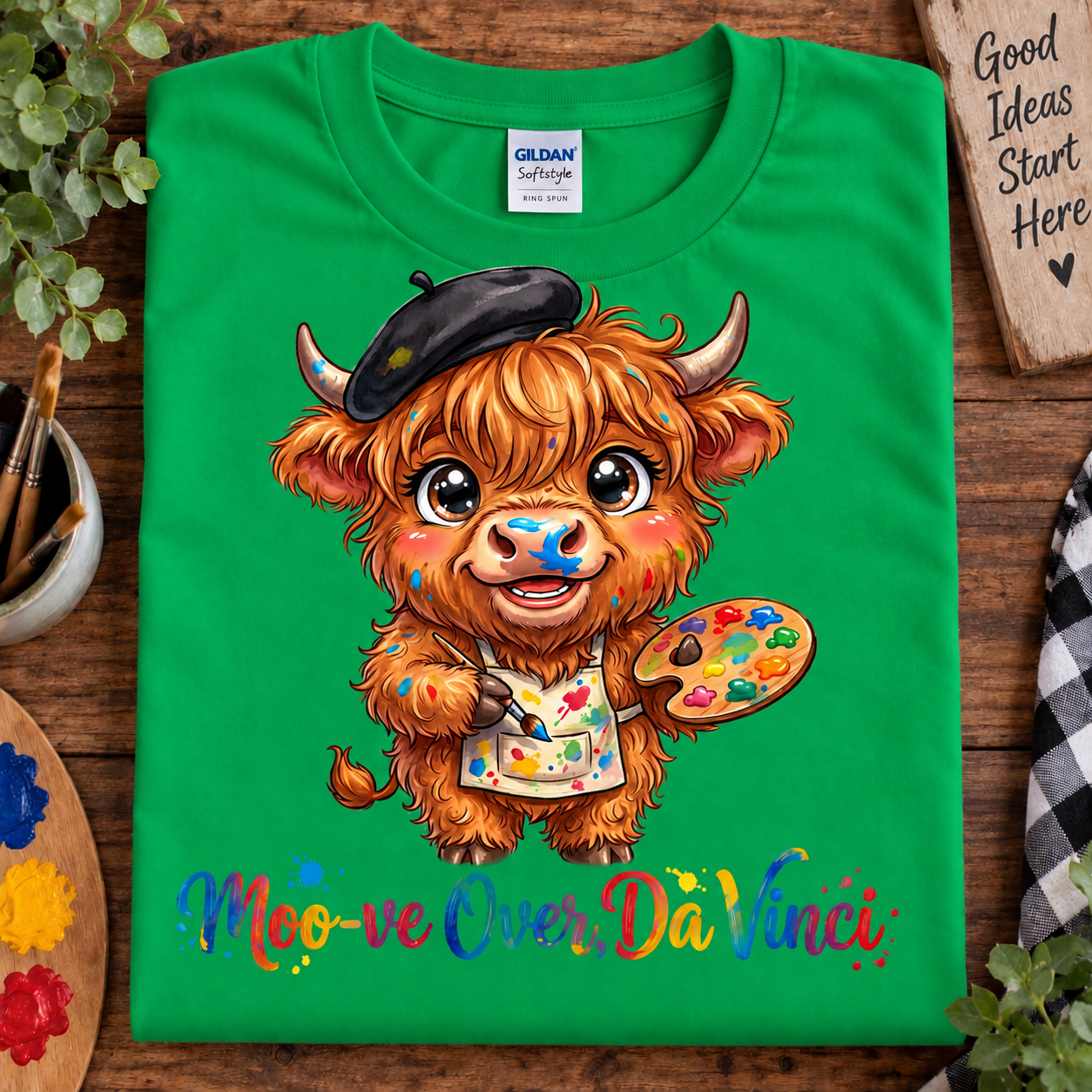 A bright irish green Gildan Softstyle t-shirt laid flat on a rustic wooden surface, featuring a cartoon illustration of a fluffy orange Scottish Highland cow wearing a black beret and paint apron, holding a paint palette. Below the illustration is the colorful text 'Moo-move Over Da Vinci'.