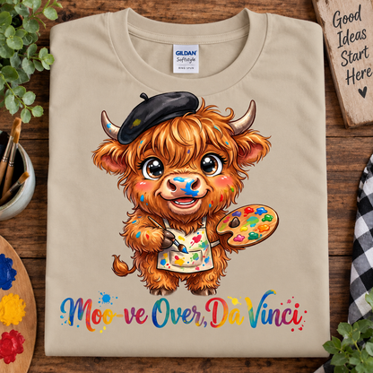 A sand Gildan Softstyle t-shirt laid flat on a rustic wooden table, featuring a cartoon illustration of a cute Highland cow dressed as Da Vinci with a black beret, paintbrush, and a paint palette. Below the illustration is the text 'Moo-ve Over, Da Vinci' in a colorful script.