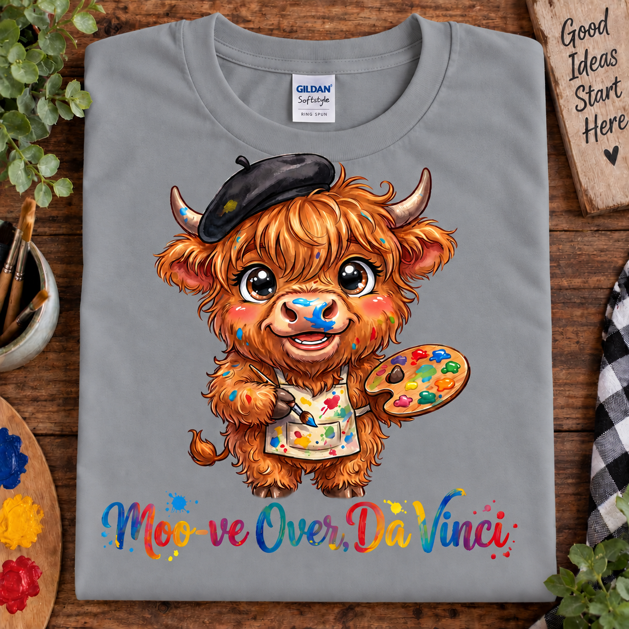 A folded sport gray Gildan t-shirt with a colorful cartoon illustration of a Scottish highland cow dressed as Da Vinci, holding a paint palette and wearing a beret. Below the illustration is the text 'Moo ve Over, Da Vinci' in a playful multicolor font.
