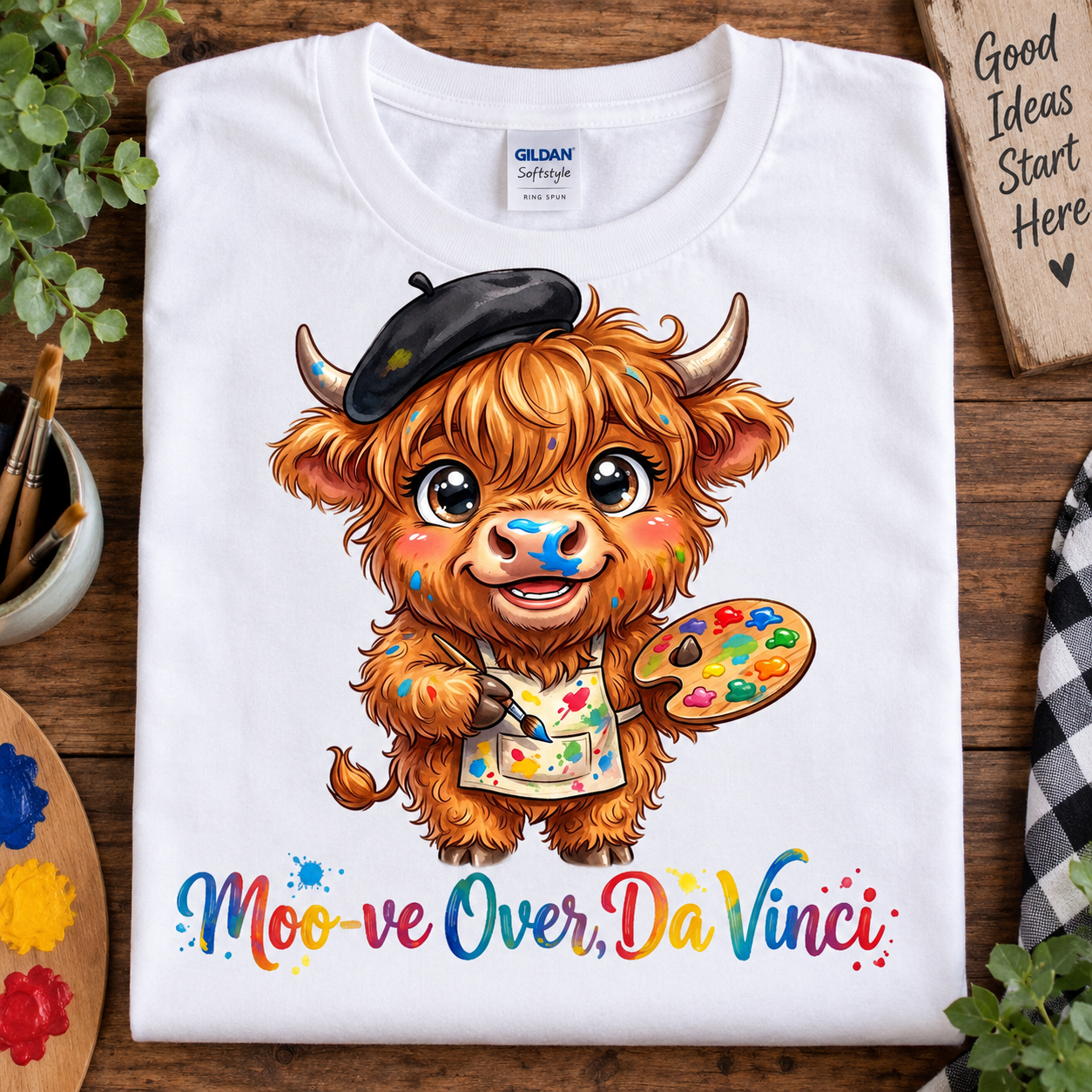 A white Gildan Softstyle T-shirt laid flat on a wooden surface, featuring a colorful cartoon illustration of a fluffy brown cow wearing a black beret and paint apron, holding a paint palette, with the text 'Moo-ve Over, Da Vinci' below. Decorative plants, paint palettes, and a wooden sign are arranged around the shirt.