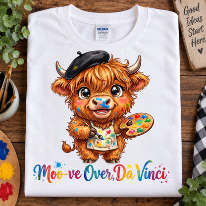 A white Gildan Softstyle T-shirt laid flat on a wooden surface, featuring a colorful cartoon illustration of a fluffy brown cow wearing a black beret and paint apron, holding a paint palette, with the text 'Moo-ve Over, Da Vinci' below. Decorative plants, paint palettes, and a wooden sign are arranged around the shirt.