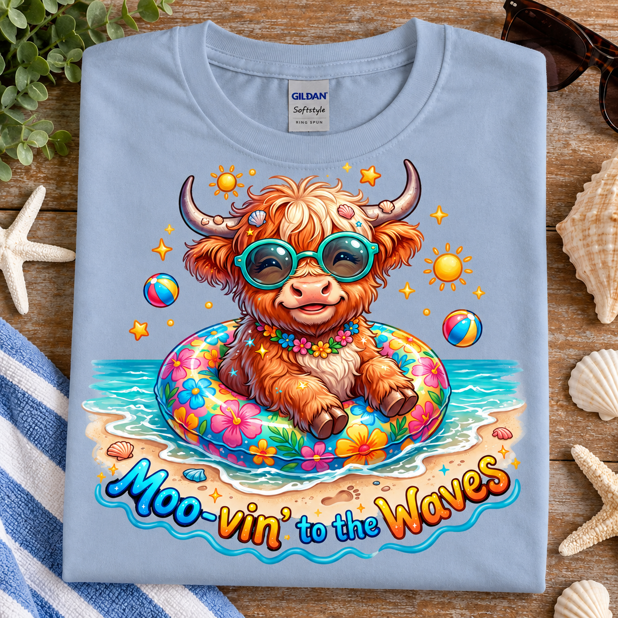 A folded light blue Gildan Softstyle T-shirt is displayed on a rustic wooden surface with beach-themed props. The shirt features a colorful graphic of a fluffy brown Highland cow sitting in a floral float on a beach, wearing teal sunglasses, with the text 'Moo-vin' to the Waves' below.