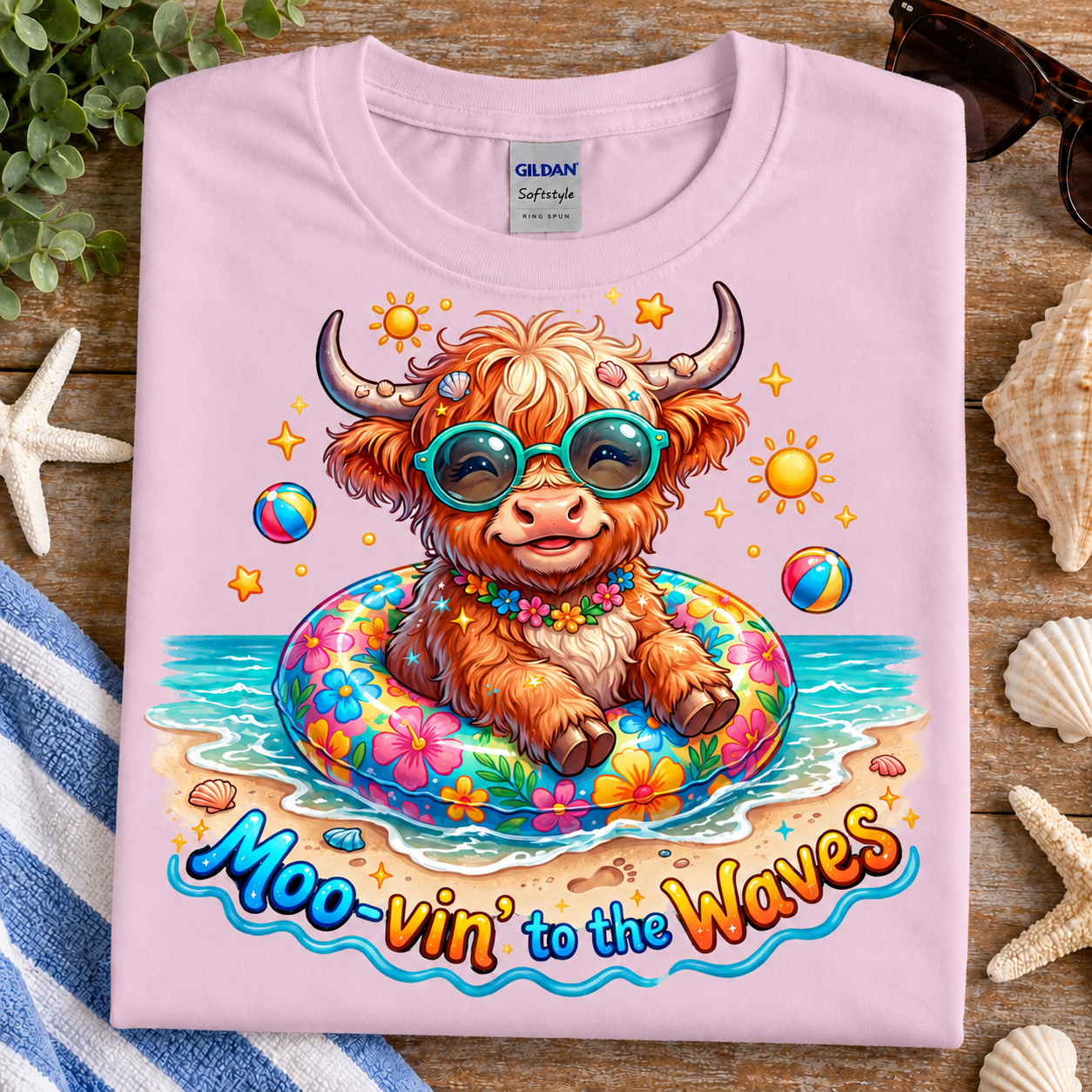 A folded light pink Gildan Softstyle T-shirt is displayed on a rustic wooden surface with beach-themed props like starfish, seashells, a towel, and sunglasses. The shirt features a colorful graphic of a fluffy Highland cow wearing teal sunglasses and sitting in a floral float on a beach, with the text 'Moo-vin' to the Waves' printed below.