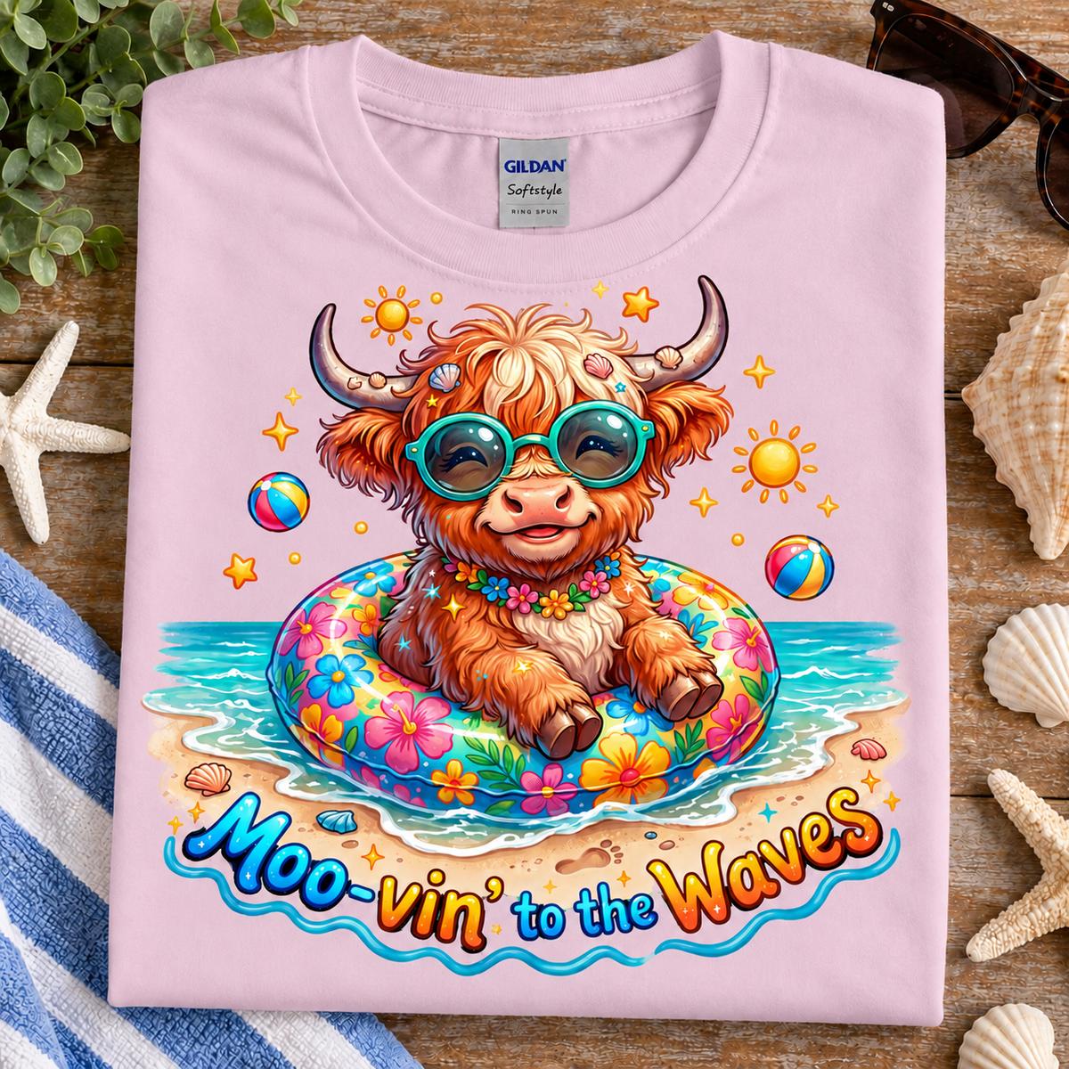 A folded light pink Gildan Softstyle T-shirt is displayed on a rustic wooden surface with beach-themed props like starfish, seashells, a towel, and sunglasses. The shirt features a colorful graphic of a fluffy Highland cow wearing teal sunglasses and sitting in a floral float on a beach, with the text 'Moo-vin' to the Waves' printed below.
