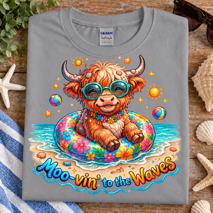 A folded sport gray Gildan Softeye t-shirt is displayed on a rustic wooden surface surrounded by beach-themed props like starfish, seashells, a blue-and-white striped towel, and sunglasses. The shirt features a colorful graphic of a fluffy Highland cow wearing teal sunglasses and sitting in a floral float on the beach, with the text 'Moo-vin' to the Waves' printed below.