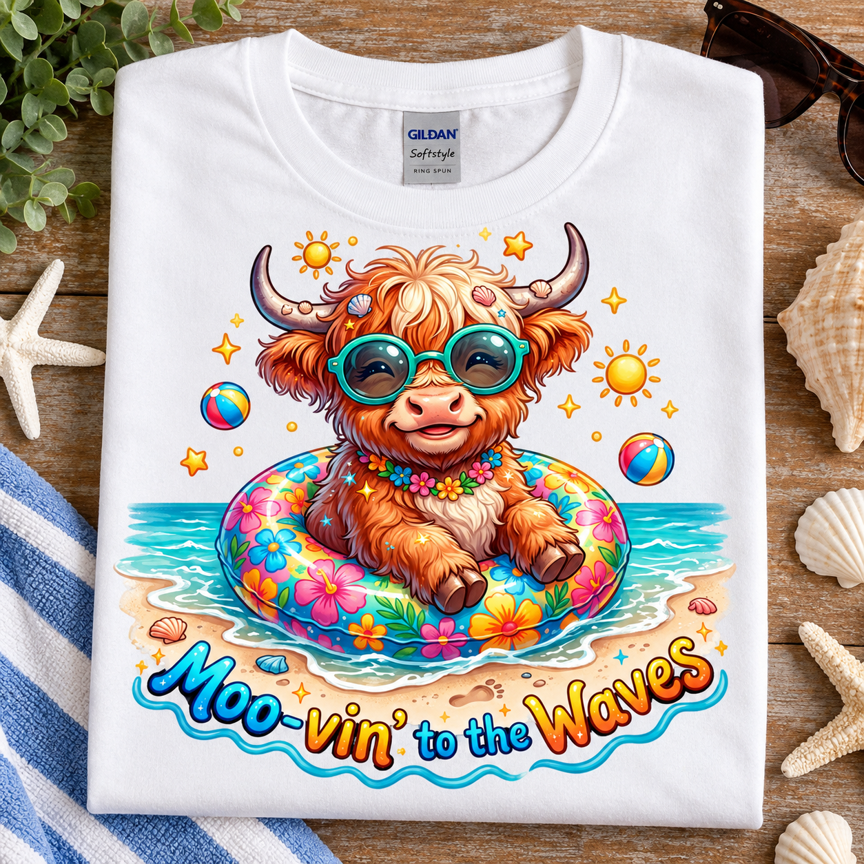 A folded white Gildan Sofstyle T-shirt with a colorful cartoon Highland cow graphic sitting in a pool float on a beach, surrounded by seashells, starfish, and a striped towel on a wooden surface.