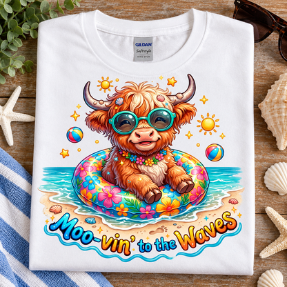 A folded white Gildan Sofstyle T-shirt with a colorful cartoon Highland cow graphic sitting in a pool float on a beach, surrounded by seashells, starfish, and a striped towel on a wooden surface.