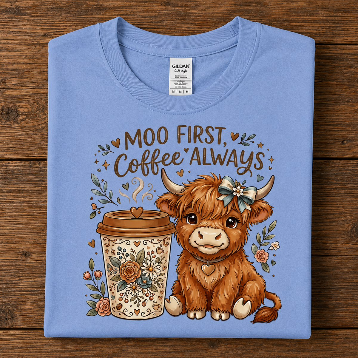 A carolina blue folded Gildan SoftSTyle  T-shirt with a graphic print of a cute brown highland cow and a coffee cup, accompanied by the text 'MOO FIRST, Coffee ALWAYS', displayed flat on a dark wood background.