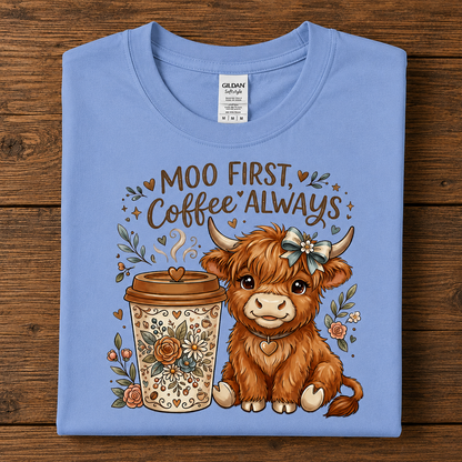 A carolina blue folded Gildan SoftSTyle  T-shirt with a graphic print of a cute brown highland cow and a coffee cup, accompanied by the text 'MOO FIRST, Coffee ALWAYS', displayed flat on a dark wood background.