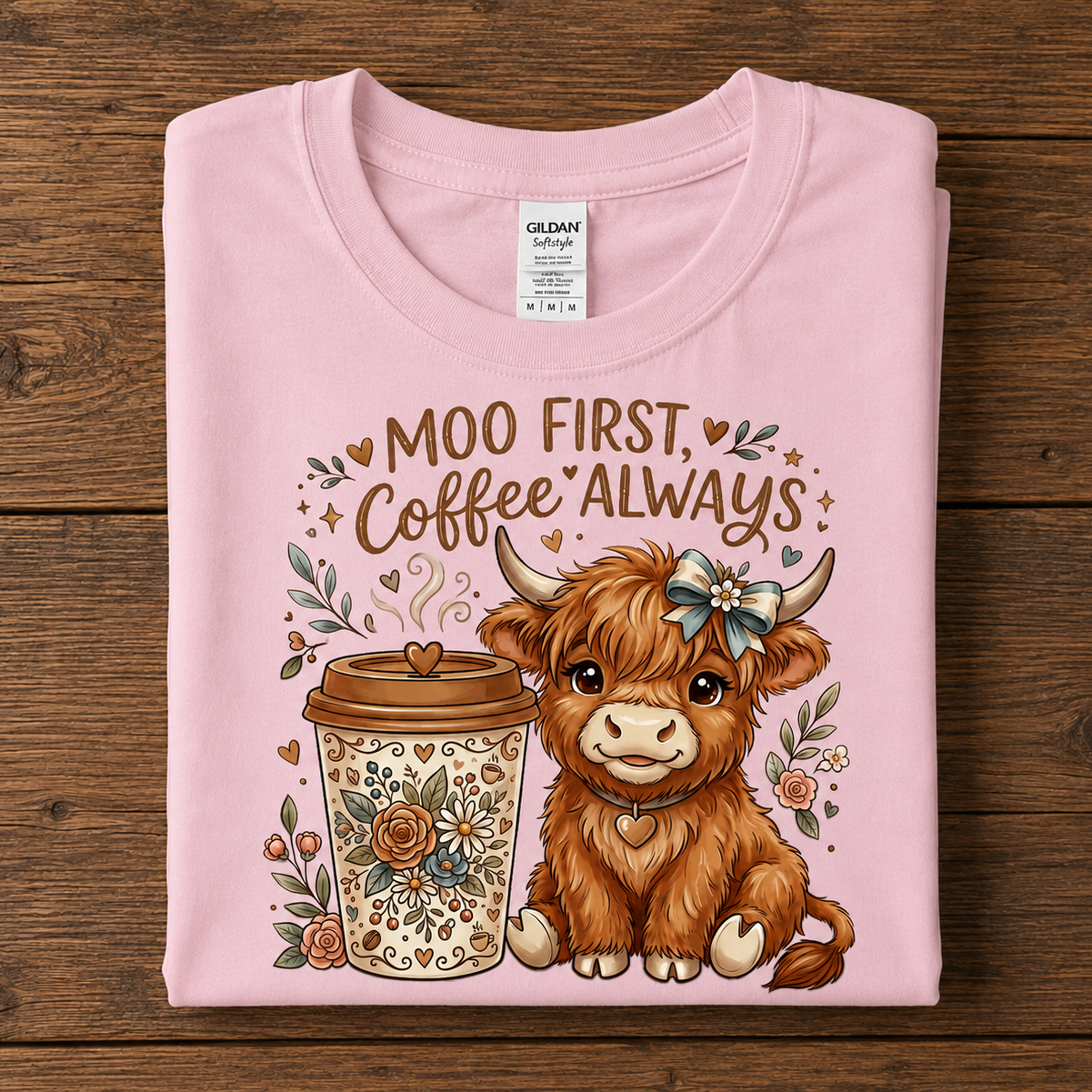 A folded light pink Gildan t-shirt with a graphic print of a cute brown Scottish highland cow and a coffee cup, accompanied by the text 'MOO FIRST Coffee ALWAYS', displayed flat on a dark wood background.