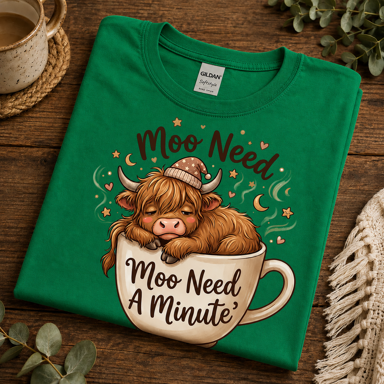 A folded turf green Gildan t-shirt with a cartoon Highland cow sleeping in a teacup and the text 'Moo Need A Minute', displayed on a rustic wooden surface with decorative props.