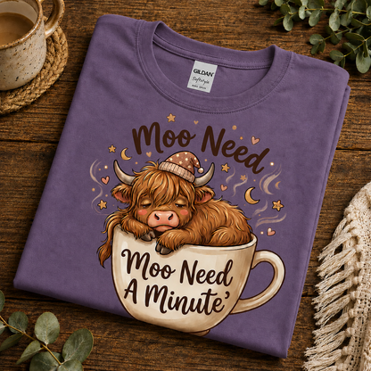 A folded heather purple Gildan Softstyle T-shirt with a cartoon highland cow sleeping in a teacup and the text 'Moo Need Moo Need A Minute' on a rustic wooden surface with decorative props.