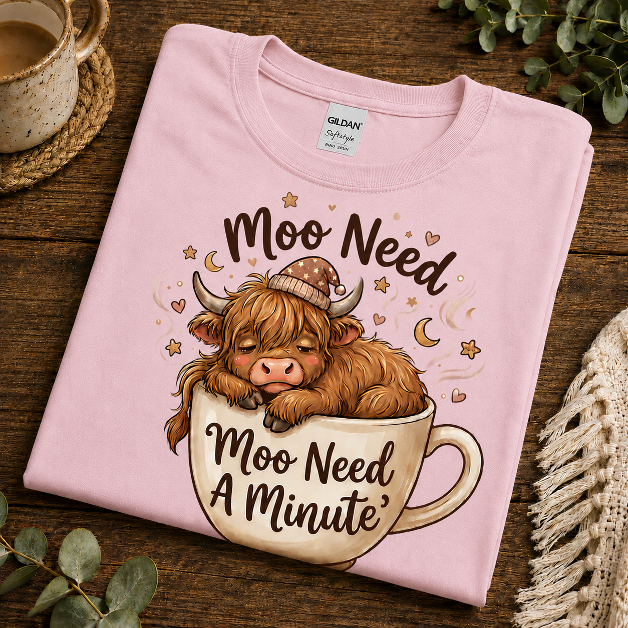 A folded light pink Gildan Softstyle T-shirt with a cartoon Highland cow sleeping in a teacup and the text 'Moo Need A Minute,' styled on a wooden surface with a mug, greenery, and a knit textile nearby.