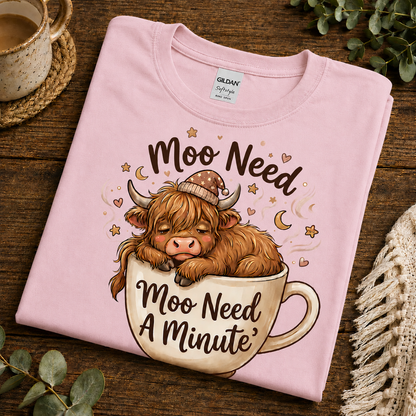 A folded light pink Gildan Softstyle T-shirt with a cartoon Highland cow sleeping in a teacup and the text 'Moo Need A Minute,' styled on a wooden surface with a mug, greenery, and a knit textile nearby.