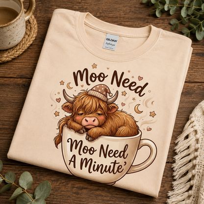 A folded natural colored Gildan Softstyle T-shirt with a cute graphic of a fluffy brown cow sleeping in a coffee mug, surrounded by stars and hearts, with the text 'Moo Need A Minute'. The shirt is displayed on a rustic wooden table with cozy lifestyle props like a ceramic mug, eucalyptus leaves, and a knit blanket.