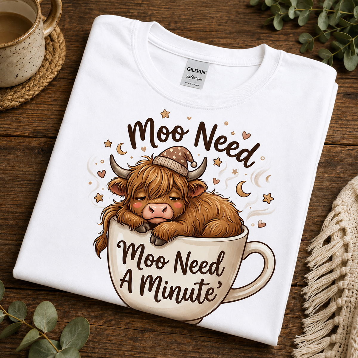A folded white Gildan Comfortware t-shirt with a cartoon illustration of a Scottish highland cow resting in a coffee mug, surrounded by the text 'Moo Need A Minute'. The shirt is displayed on a rustic wooden table with cozy lifestyle props like a mug, eucalyptus leaves, and a knit blanket.