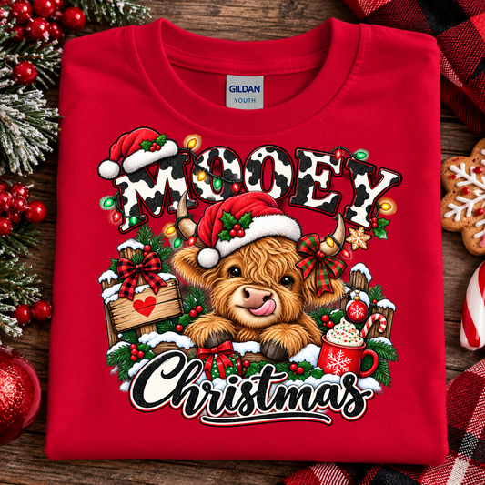 A folded red Gildan Youth t-shirt with a festive 'Mooey Christmas' graphic, styled on a rustic wooden background with holiday decorations like pine branches, red berries, gingerbread cookies, and a plaid blanket.