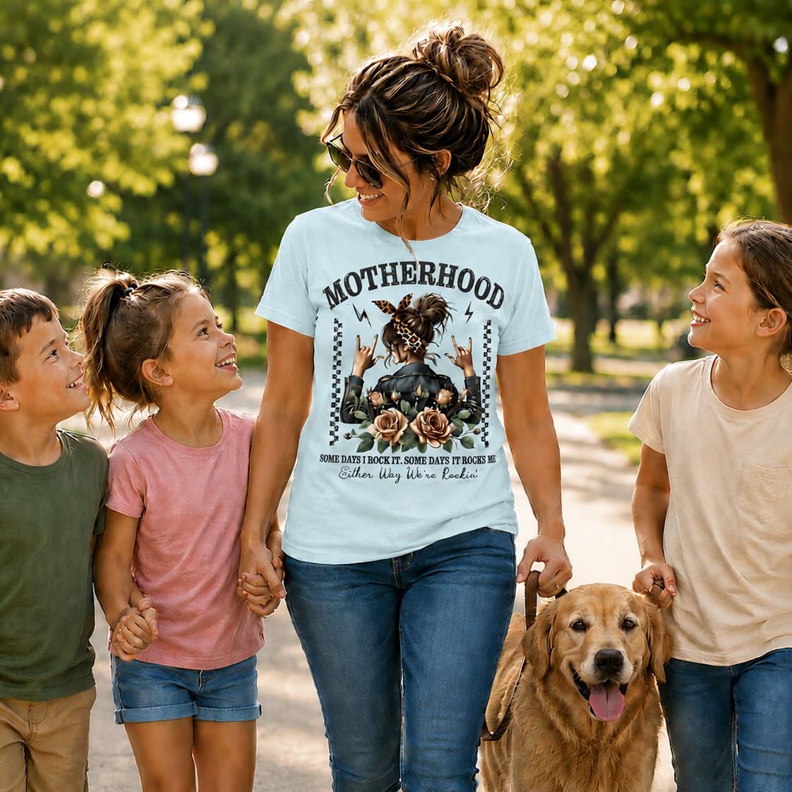 Woman wearing a chambray colored(blue) graphic t-shirt with Motherhood saying on it walking with three children and a dog in a park