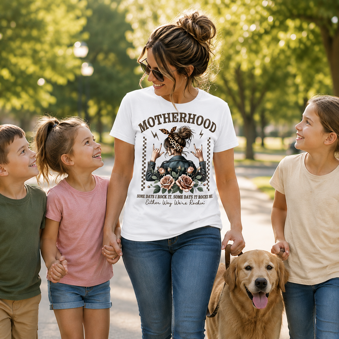 Woman wearing a white graphic t-shirt with Motherhood saying on it walking with three children and a dog in a park