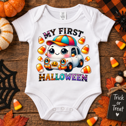 A white short-sleeve baby bodysuit laid flat on a rustic wooden surface, decorated with Halloween-themed props including pumpkins, candy corn, autumn leaves, a spiderweb cutout, and a small sign reading 'Trick or Treat.' The bodysuit features a colorful cartoon graphic of a smiling car with the text 'MY FIRST HALLOWEEN.