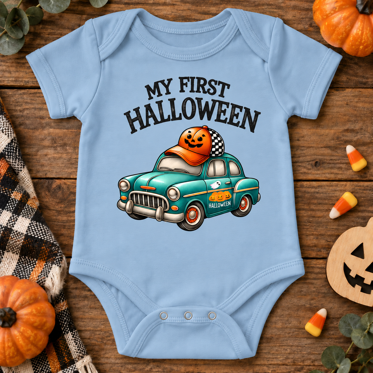 A light blue baby bodysuit with a 'My First Halloween' graphic featuring a teal car and a jack-o'-lantern, styled in a flat lay on a wooden surface with seasonal props.