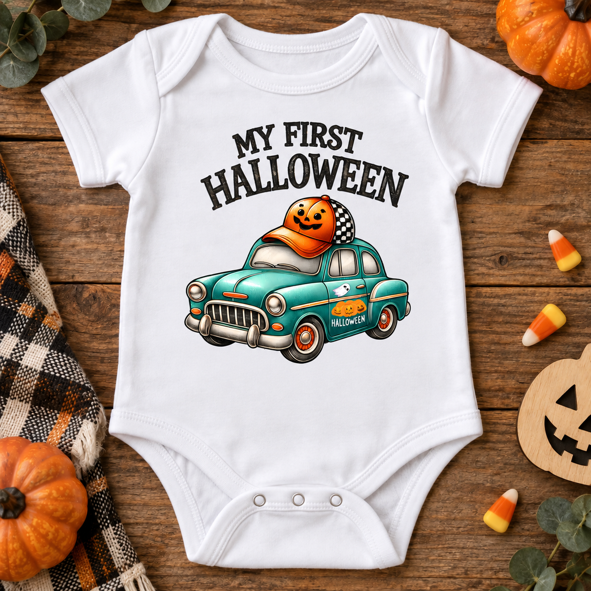 A white short-sleeve baby bodysuit laid flat on a rustic wooden surface, printed with the phrase 'MY FIRST HALLOWEEN' above a teal car carrying a jack-o'-lantern. The scene is styled with small pumpkins, candy corn, a plaid cloth, and a wooden jack-o'-lantern cutout.