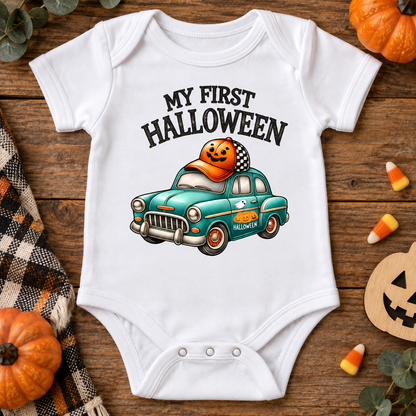 A white short-sleeve baby bodysuit laid flat on a rustic wooden surface, printed with the phrase 'MY FIRST HALLOWEEN' above a teal car carrying a jack-o'-lantern. The scene is styled with small pumpkins, candy corn, a plaid cloth, and a wooden jack-o'-lantern cutout.