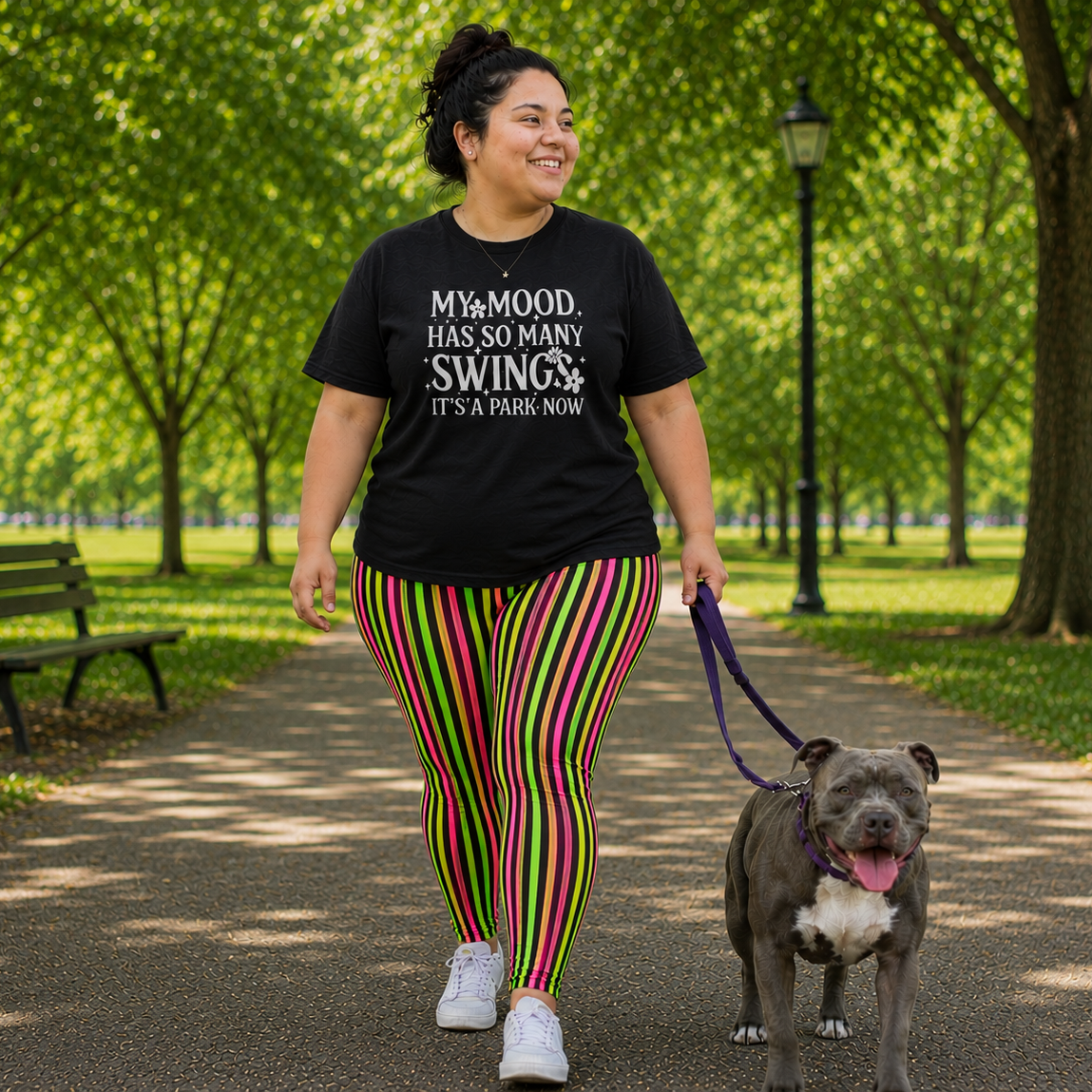 Person walking a dog in a park wearing a black t-shirt with text and colorful pants.