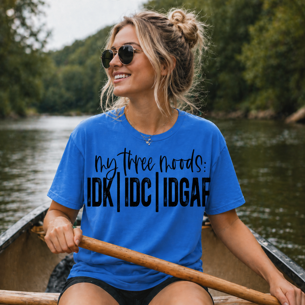 Woman in a heather royal t-shirt with humorous text on a canoe, surrounded by nature.