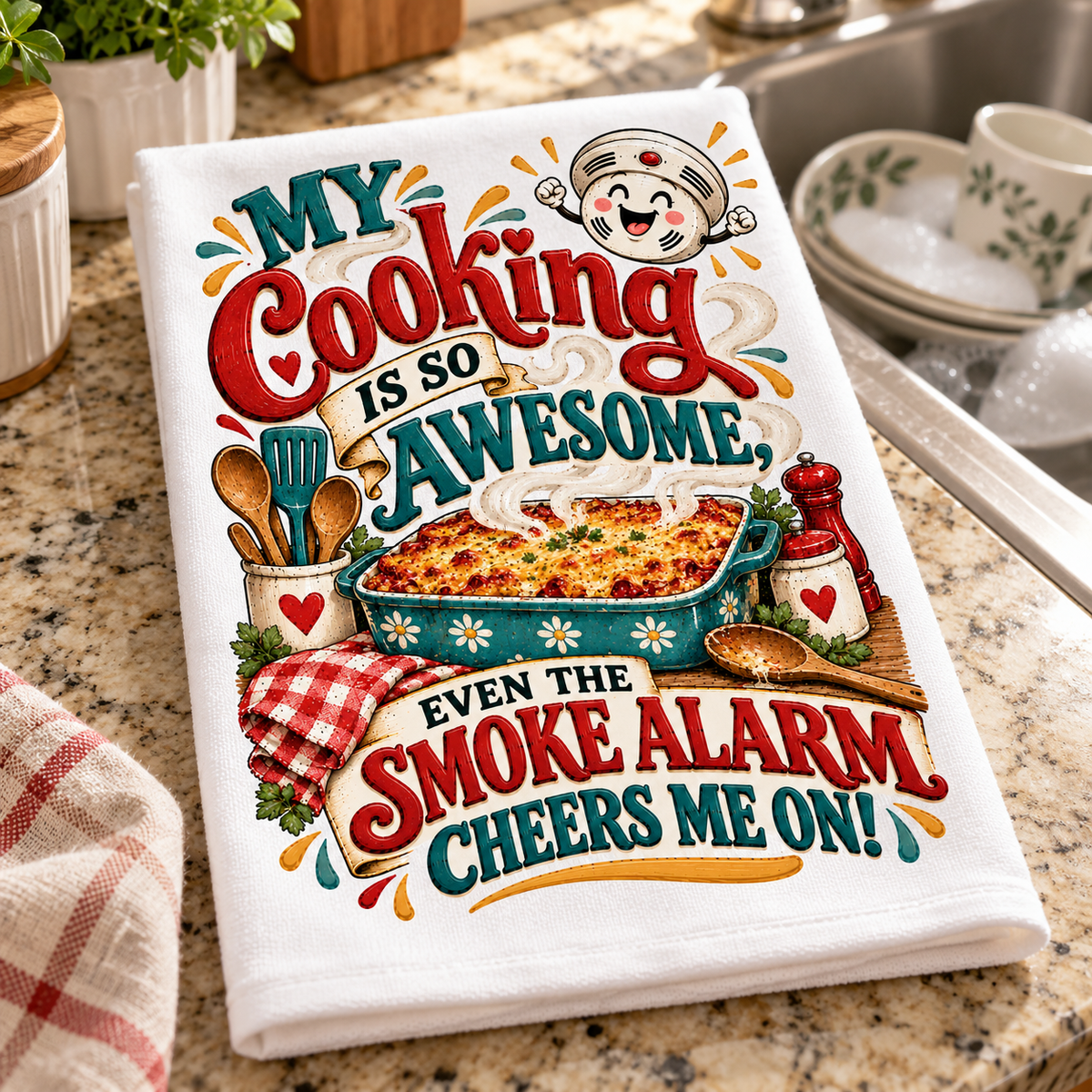 A white kitchen towel with a colorful retro-style cooking graphic and the text 'MY Cooking IS SO AWESOME, EVEN THE SMOKE ALARM CHEERS ME ON!' is laid on a granite countertop near a sink with dishes.