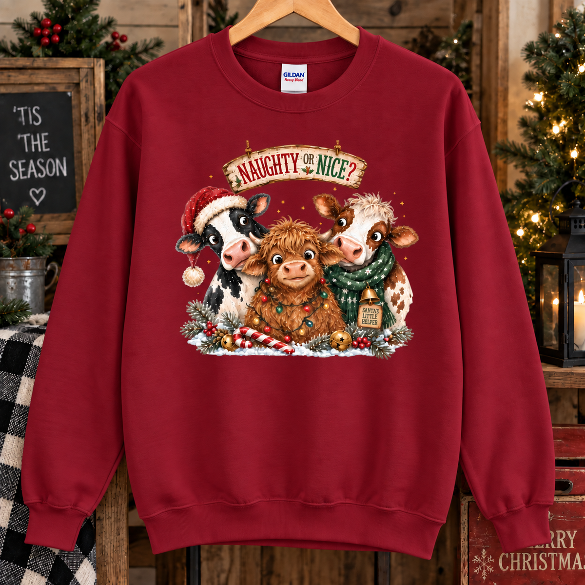 A antique cherry red crewneck sweatshirt hanging on a wooden hanger, featuring a festive graphic of three cute cows dressed in Christmas attire with the text 'NAUGHTY NICE' above them. The background is a cozy, rustic Christmas setting with a lit tree, candles, and holiday decorations.