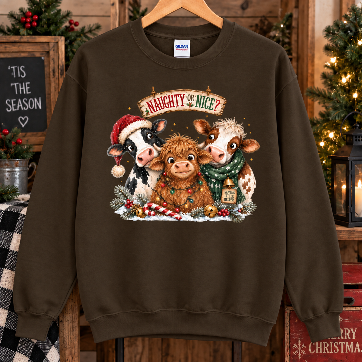 A dark chocolate brown crewneck sweatshirt hanging on a wooden hanger, featuring a festive Christmas graphic of three cows with Santa hats and scarves under the text 'Naughty Nice?'. The background is a rustic holiday scene with Christmas trees, candles, and wooden decor.