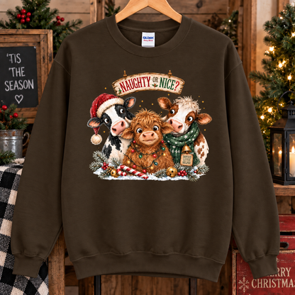A dark chocolate brown crewneck sweatshirt hanging on a wooden hanger, featuring a festive Christmas graphic of three cows with Santa hats and scarves under the text 'Naughty Nice?'. The background is a rustic holiday scene with Christmas trees, candles, and wooden decor.