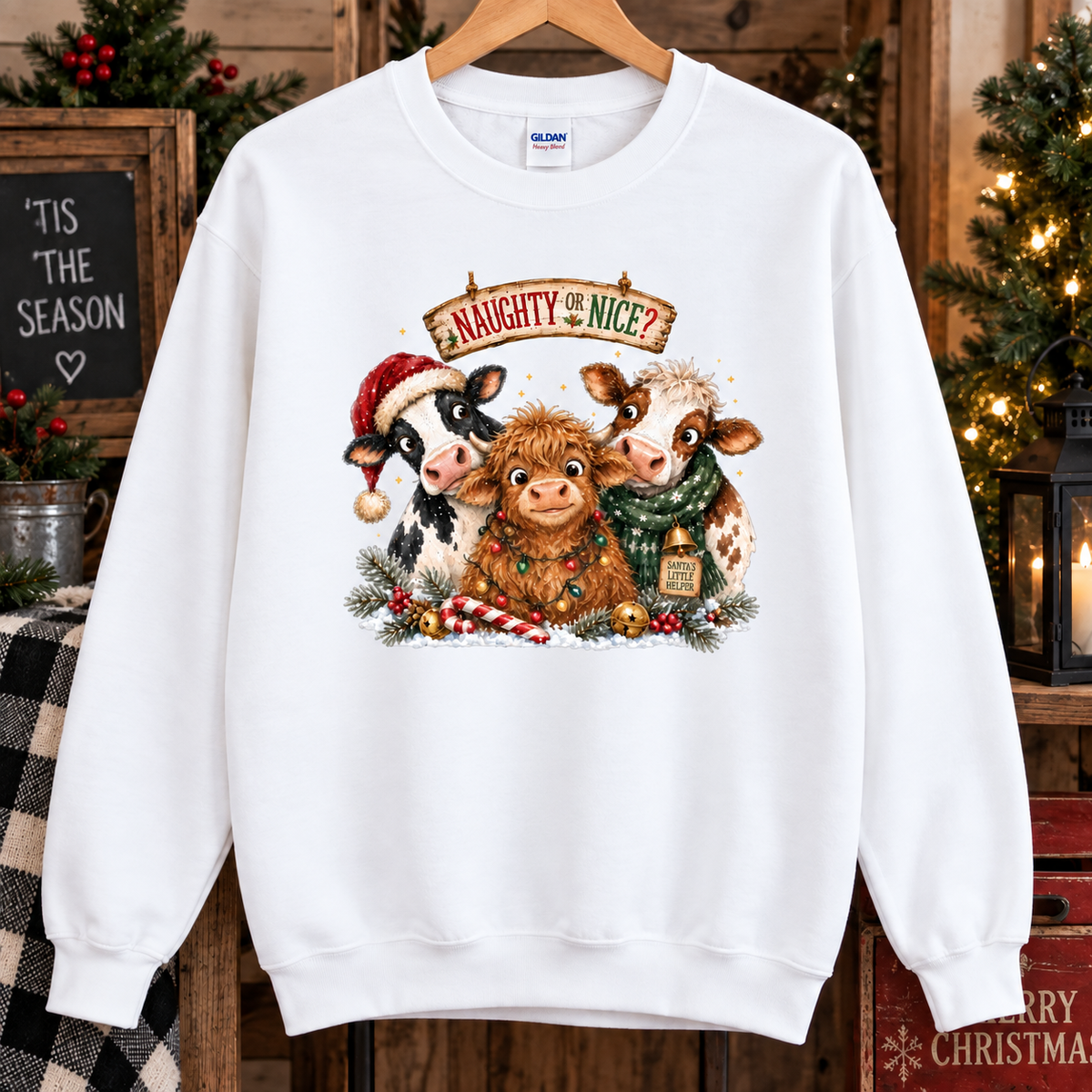 A white crewneck sweatshirt hanging on a wooden hanger, featuring a festive graphic of three cute cows dressed in Christmas attire with the text 'A NAUGHTY NICE?' above them. The background is a cozy, rustic holiday scene with Christmas trees, candles, and wooden decorations.