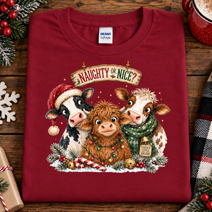 A folded antique cherry red Gildan Softstyle t-shirt is displayed on a rustic wooden background with Christmas-themed props like pine branches, berries, a mug, and a plaid scarf. The shirt features a graphic of three cute cows and a Highland cow decorated for Christmas, with the text 'NAUGHTY OR NICE?'.