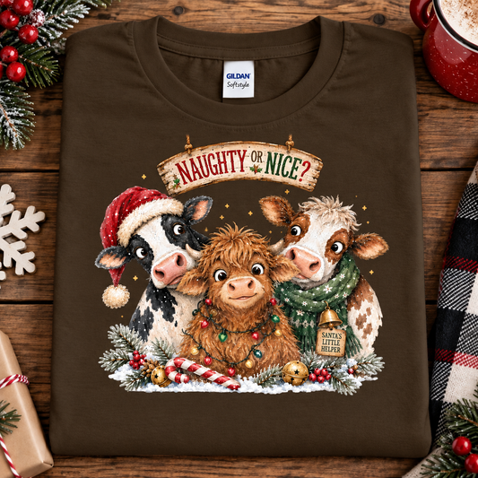 A dark chocolate brown Gildan Softstyle t-shirt laid flat on a rustic wooden table, featuring a festive Christmas graphic of three cows and a Highland cow with holiday hats and scarves under a banner that reads 'NAUGHTY OR NICE?'.