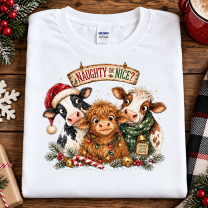 A folded white Gildan Softstyle t-shirt with a festive Christmas graphic of three cows and the text 'NAUGHTY OR NICE?'. The shirt is styled in a flat-lay on a rustic wooden table surrounded by holiday decorations like pine branches, red berries, a mug, and a plaid blanket.