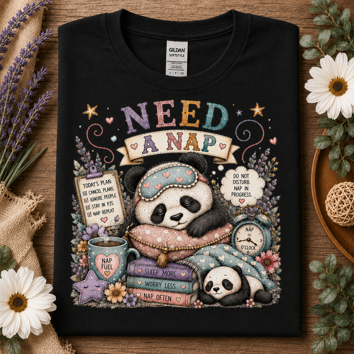 A black Gildan Softstyle t-shirt laid flat on a rustic wooden surface, surrounded by decorative props including white flowers, lavender, burlap, and a small woven ball. The shirt features a colorful graphic of two pandas sleeping under a blanket, with text reading 'NEED A NAP' and additional nap-themed illustrations.