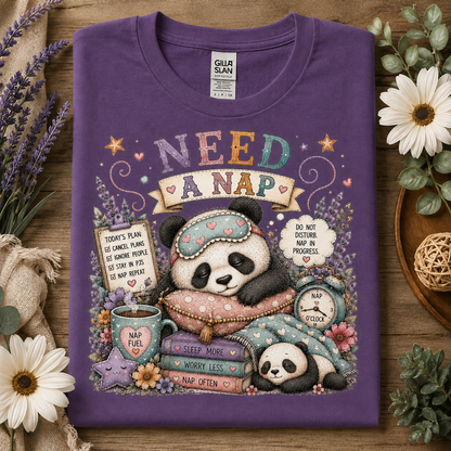 A folded heather purple crewneck t-shirt with a colorful cartoon panda nap graphic, displayed on a rustic wooden surface with flowers and greenery.