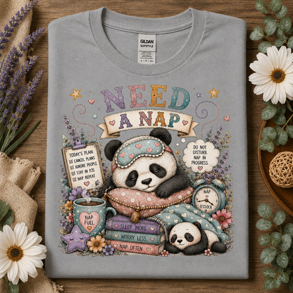 A folded sport gray Gildan Supreme t-shirt with a colorful cartoon panda nap graphic and the text 'NEED A NAP', styled on a wooden surface with flowers and burlap.