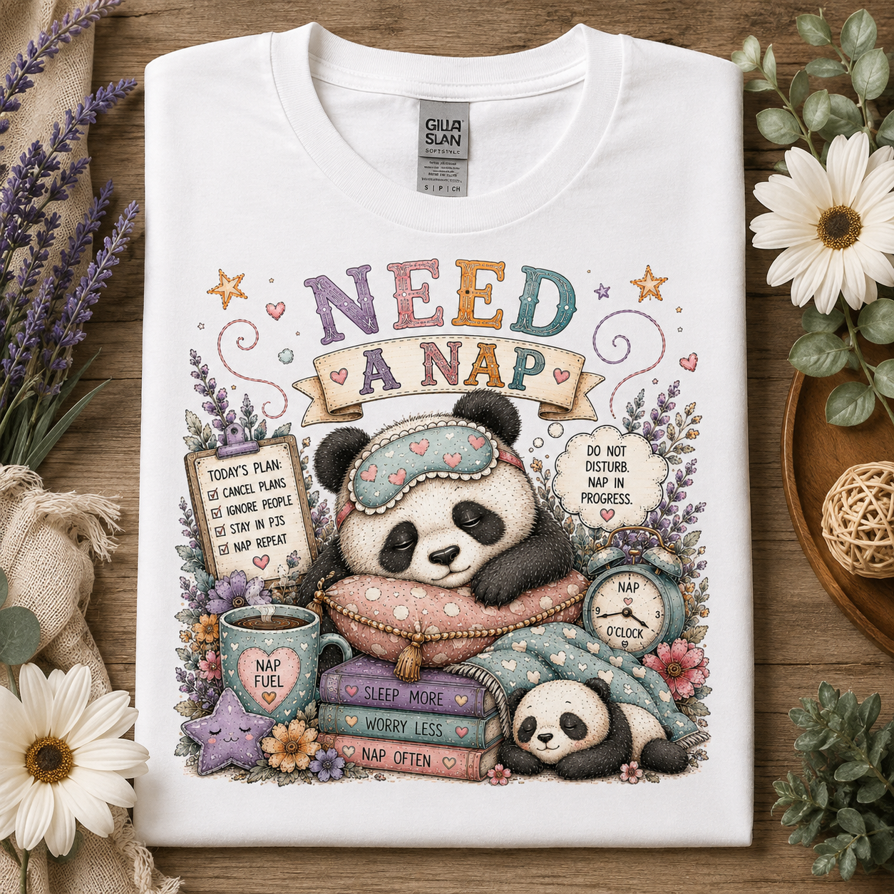 A white short-sleeve T-shirt laid flat on a wooden surface, surrounded by decorative flowers, lavender, and burlap. The shirt features a large colorful panda nap graphic with the words “NEED A NAP,” a mug reading “NAP FUEL,” books, stars, and small text bubbles.