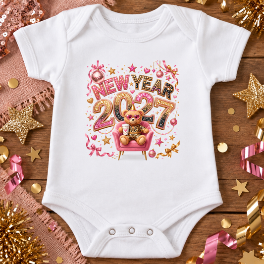 A white short-sleeve baby bodysuit laid flat on a rustic wooden surface, decorated with a colorful 'New Year 2027' graphic featuring a teddy bear in a pink chair. The bodysuit is surrounded by festive gold and pink tinsel, stars, and sequins.