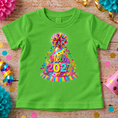 A apple green short-sleeve kids' t-shirt laid flat on a rustic wooden surface, decorated with colorful party-themed props. The front of the shirt features a large, vibrant graphic of a party hat with a pom-pom, filled with confetti and the words 'Hello 2021' in a rainbow-colored script.